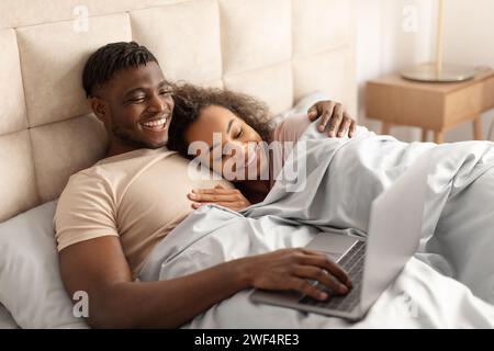 Happy black man and woman using laptop lying in bed Stock Photo