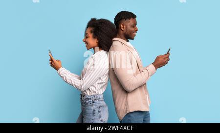 Black man and woman using smartphones back-to-back on blue backdrop Stock Photo