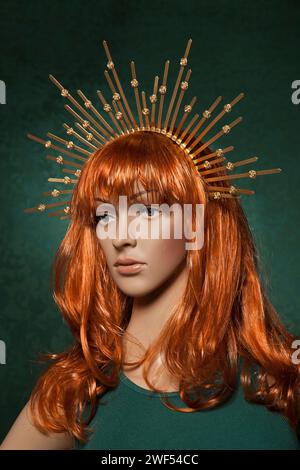 Plastic red haired woman mannequin with bright long hair wearing a spiky golden crown posing on a green background in a green tank top Stock Photo