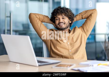 Young indian man in formal clothes stretching back while sitting by desktop and looking at laptop screen with smile. Confident employee feeling no stress and watching result of successful working day. Stock Photo