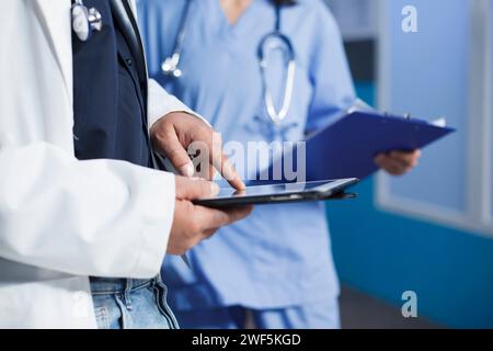 Doctor using a digital tablet in medical office while wearing a lab coat. Nurse waits with clipboard in background with selective focus on masculine hands grasping a smart gadget. Stock Photo