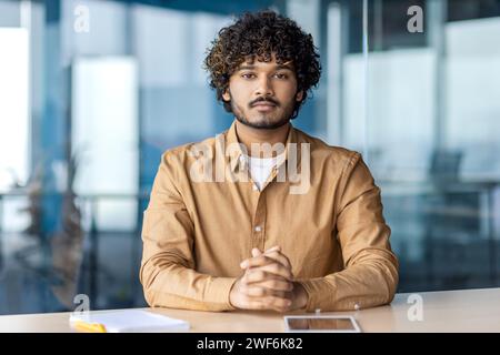 Strict indian businessman sitting with hands folded on table and looking confidently at camera on blurred background. Focused male thinking over company future and pondering over project development. Stock Photo