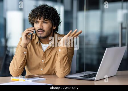 Serious arab man having unpleasant phone call during working day in modern office interior. Annoyed young male contacting customer support about faulty laptop and getting unsatisfactory answer. Stock Photo
