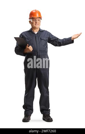 Male factory worker in an overall uniform and orange helmet holding a clipboard and pointing isolated on white background Stock Photo