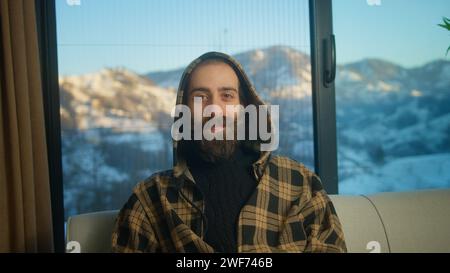 Bearded young man wearing hoodie relaxing, looking at camera and smiling with snowy mountain background Stock Photo
