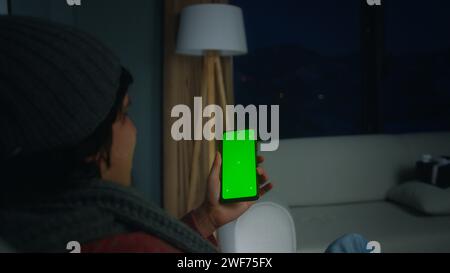 Young man using, tapping on screen smartphone with green mock-up screen. Male sit on armchair, relaxing at home. Close-up over the shoulder shot Stock Photo