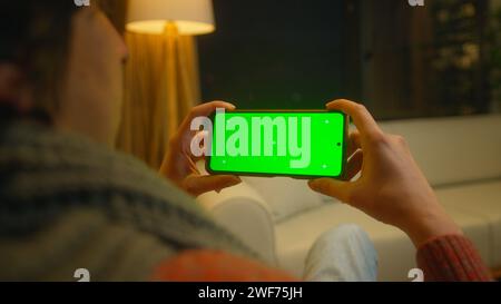 Young man watching video on his smartphone with green screen mock up display, which he holds horizontally. Male sitting on sofa relaxing at home. Clos Stock Photo