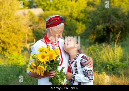 Gray haired grandmother and girl granddaughter both wearing embroidered shirts with a bouquet of sunflowers smiling snd happy. Independence day, const Stock Photo