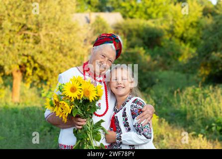 Gray haired grandmother and girl granddaughter both wearing embroidered shirts with a bouquet of sunflowers smiling snd happy. Independence day, const Stock Photo