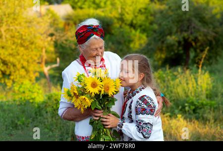 Gray haired grandmother and girl granddaughter both wearing embroidered shirts with a bouquet of sunflowers smiling snd happy. Independence day, const Stock Photo