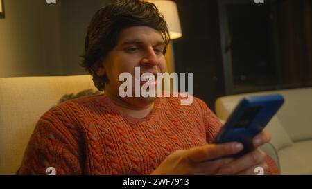 Cosy man sit on armchair in living room using smartphone browses through internet, watches videos and uses social networks Stock Photo
