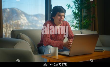Young man wear wireless headset with microphone, looking at laptop, having video conference call talk by webcam while sittin on sofa in living room Stock Photo