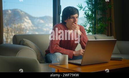 Distracted young student working from home using with laptop while sitting on sofa in living room. Bored man student trying to study, staring blankly Stock Photo