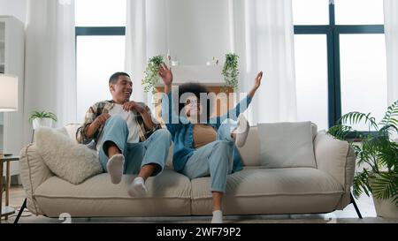 Happy African American ethnic couple jumping on sofa laugh having fun fooling around enjoy relax rest fall on cozy couch celebrate moving day Stock Photo
