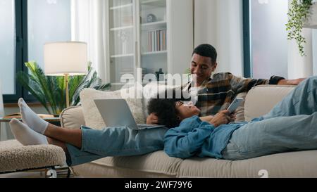 Chilling African American relaxed couple on couch at home together gadget internet addict man working using laptop woman laying browsing mobile phone Stock Photo