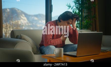 Young man sitting on sofa suffering chest pain, having heart attack, holding massaging chest while working with laptop. Male trouble breathing stroke Stock Photo