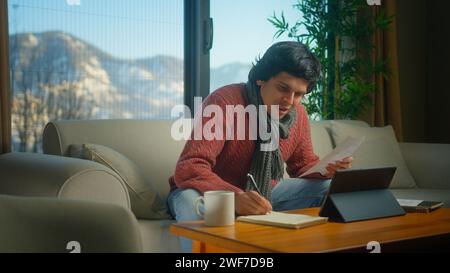 Young student wear scarf sit on sofa doing research, homework with tablet device, taking notes. Man peaceful studying at home Stock Photo