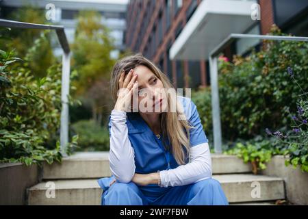 Young female doctor feeling overwhelmed at work, sitting on stairs, looking at phone. Healthcare workers having stressful job, feeling exhausted Stock Photo