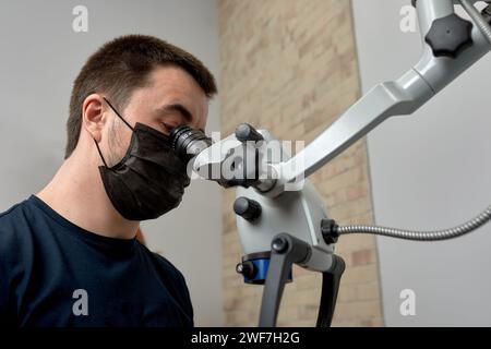 Male dentist looks through a dental microscope to examine patient's teeth. Doctor in a surgical mask uses high-tech dental equipment in his daily work Stock Photo