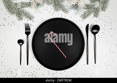 Christmas composition, black plate and cutlery, fir tree branch, candy cane, silver stars confetti on white background. Stock Photo