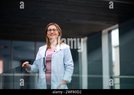 Portrait of female doctor standing in hospital, leaning on glass railing, looking at camera. Stock Photo