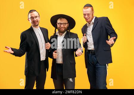 Three Jewish men, friends in traditional Jewish attributes smiling, posing against yellow studio background Stock Photo