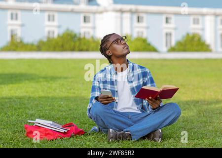 Thoughtful black guy reading book in park and using smartphone Stock Photo
