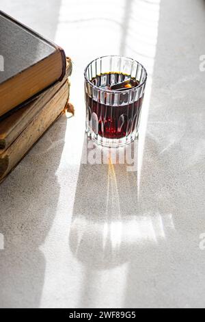 A faceted glass of cherry liqueur casting a long shadow near a pile of old books in sunlight Stock Photo