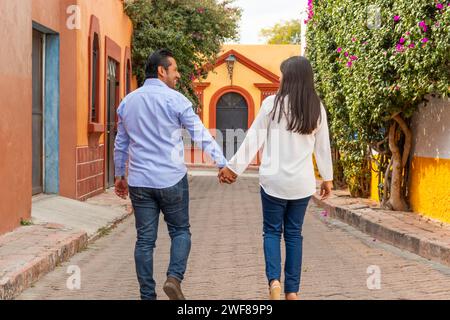 Back view of couple walks hand in hand along a colorful street in Tequisquiapan Queretaro Mexico surrounded by vibrant walls and blooming flowers Stock Photo