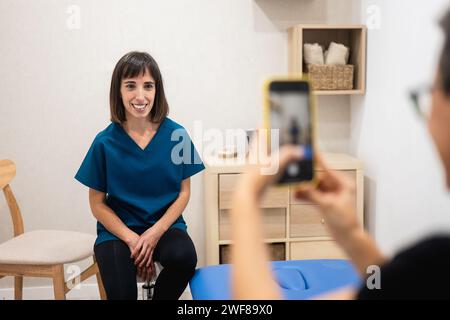 A cheerful female healthcare worker in scrubs poses for a photo, with her image captured on a smartphone by another person Stock Photo