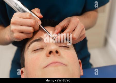A close-up shot capturing a professional grooming a client's eyebrows using stainless steel tweezers in a beauty salon Stock Photo