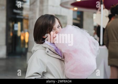 A girl with a beige jacket looking at camera while eating a large pink cotton candy on a city street Stock Photo