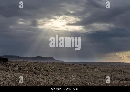 Dramatic crepuscular rays break through stormy clouds over the rocky terrain of Fuerteventura north coast, between El Cotillo and Majanicho Stock Photo