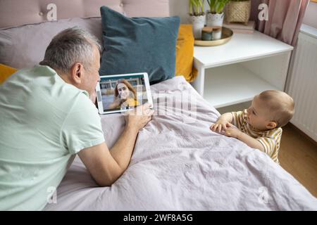 Grandfather and baby video calling a young woman from a tablet in a cozy bedroom Stock Photo