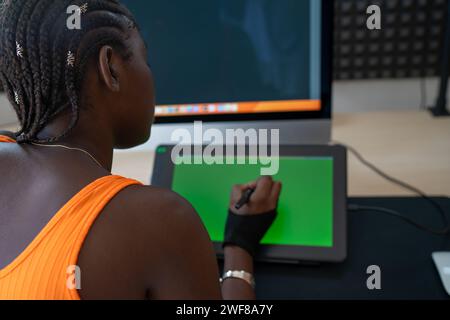 Back view of crop black lady writing with stylus pen on tablet screen while working remotely from workplace against desktop computer on background Stock Photo