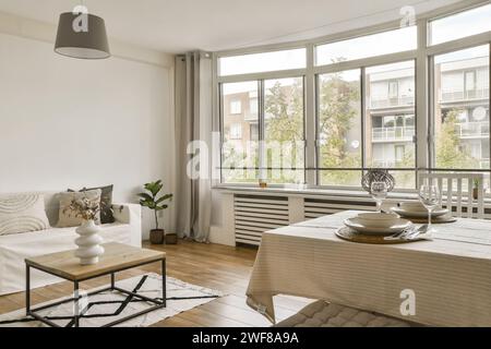 A spacious and well-lit modern living room with large windows, a cozy sofa, and a dining area set for two. Stock Photo