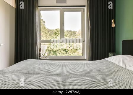 A comfortable bedroom with a patterned bedspread and dark curtains against a vibrant green accent wall, overlooking lush trees through a clear window. Stock Photo