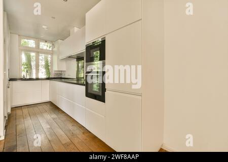 A bright and airy kitchen space featuring sleek white cabinetry, built-in appliances, and rustic wooden flooring with a view of greenery through the w Stock Photo