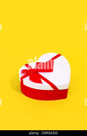 A white heart shaped gift box with a red ribbon on a vibrant yellow background Stock Photo