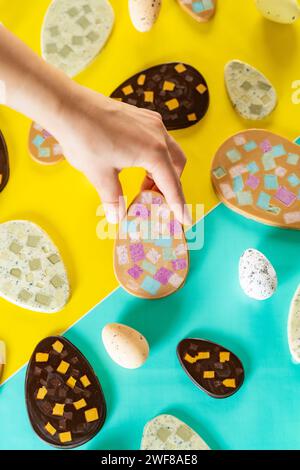 From above cropped unrecognizable person holding decorated cut in half Easter eggs scattered on a two-tone backdrop with a person reaching for one. Stock Photo