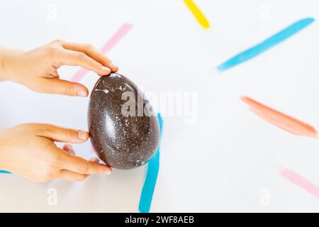 Cropped unrecognizable person's hand gently clasps a large speckled decorative egg against a white surface with vibrant paint strokes. Stock Photo
