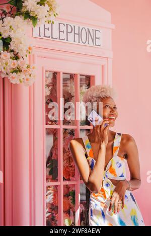 Positive young panamanian woman in stylish outfit smiling and talking on cellphone while standing near pink window in cozy room Stock Photo