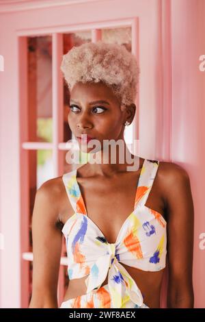 Confident panamanian woman in stylish outfit looking away while standing near window in light pink room Stock Photo