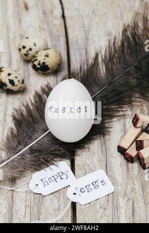 A white egg with "easter" stamped on it, surrounded by quail eggs, feathers, and "Happy eggs" tags on wooden surface. Stock Photo