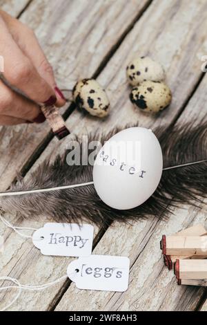 A person stamps the word 'Easter' onto a white egg, with quail eggs, feathers and tags with 'Happy' and 'eggs' written on them, laid on rustic wood. Stock Photo