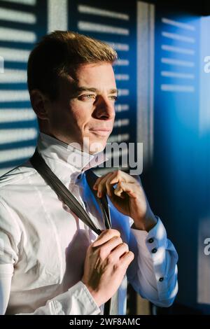 An elegant, well-groomed man with a thoughtful expression stands by a window in Madrid, adjusting his tie in the serene sunlight filtering through bli Stock Photo