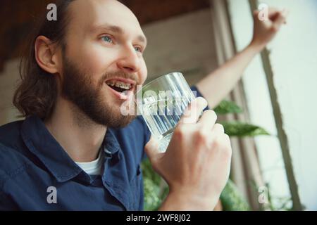 Close up photo of young bearded man in dental braces system drinking water against blurred background with plants. Stock Photo