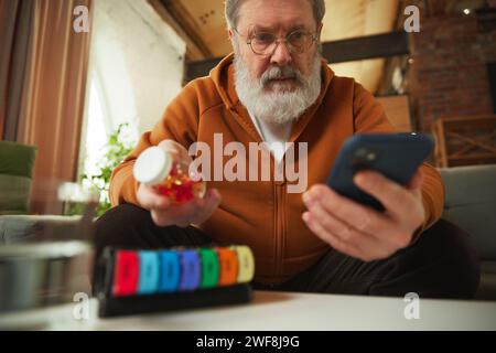 Elderly man examining pill bottle while looking at smartphone. Using pill organizer. Online med services Stock Photo