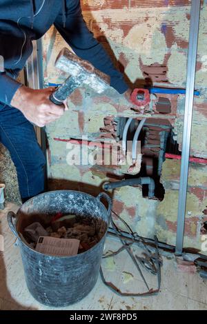 Bricklayer chipping a brick wall at a construction site to make way for the water pipes with a basket of debris. Home renovation concept Stock Photo