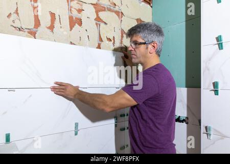 Tiler with protective glasses placing a piece of large format tile on a brick wall in a bathroom renovation. Home renovation concept Stock Photo
